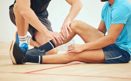 Unknown athletic squash player helping friend with injury after playing game on court. Mixed race athlete suffering from shin splints during training practice at sports centre. Physio massage for pai.