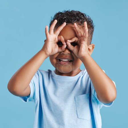Photo pour Portrait de curieux petit garçon curieux regardant à travers les doigts en forme de jumelles sur un fond de studio bleu. Enfant curieux explorer. - image libre de droit