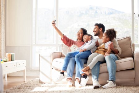 Téléchargez les photos : Selfie de famille d'une femme afro-américaine, son mari et ses filles assis sur le canapé dans le salon à la maison. Une jeune femme prenant des photos avec sa heureuse famille métissée dans le salon. - en image libre de droit