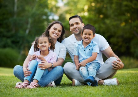 Young happy mixed race family relaxing and sitting on grass in a park together. Loving parents spending time with their little children in a garden. Carefree siblings bonding with their mom and dad.