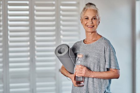 Photo for This is what I do every morning. Portrait of a cheerful mature woman holding a bottle of water and yoga mat ready to start her morning session of yoga inside of a studio - Royalty Free Image