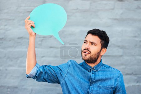 Foto de ¿Estás seguro de eso? un joven guapo que se ve escéptico mientras sostiene una burbuja del habla contra una pared de ladrillo - Imagen libre de derechos