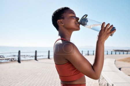 Black woman, drinking water or bottle in fitness workout, training or exercise by beach, ocean or sea in summer location. Smile, happy or sports runner with drink for healthcare wellness recovery.