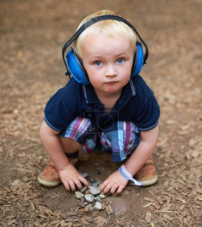 Foto de Manteniéndose ocupado en la naturaleza. un niño pequeño sentado y mirando a la cámara con auriculares protectores sobre sus orejas - Imagen libre de derechos