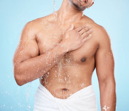 Téléchargez les photos : Body, water and shower with a man model standing in studio on a blue background for hygiene or hydration. Splash, health and wellness with a male wearing a towel in the bathroom after bathing. - en image libre de droit
