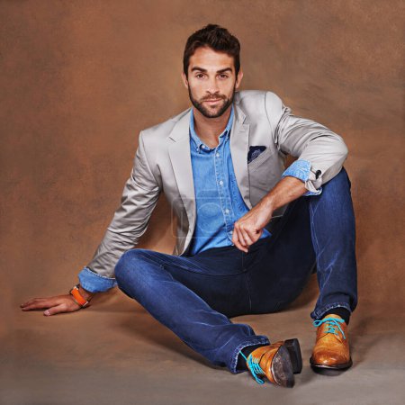 He represents the perfect example of modern elegance. a stylishly dressed young man sitting against a brown background