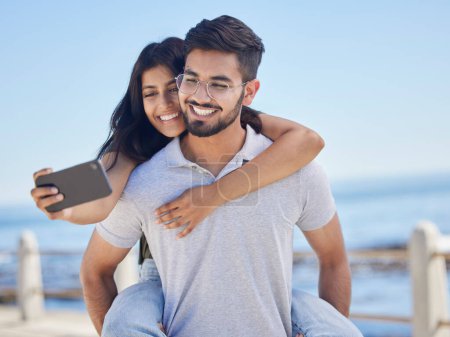 Téléchargez les photos : Phone selfie, ocean and couple hug, bond and enjoy time together for peace, freedom or romantic date. Sea beach, memory photo and man piggyback woman on fun travel holiday in Rio de Janeiro Brazil. - en image libre de droit