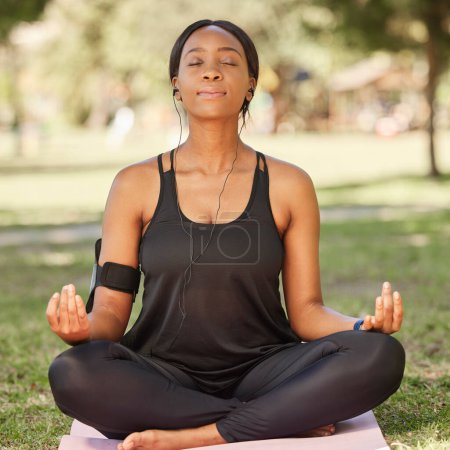 Foto de Yoga, black woman meditation in park with lotus pose, earphone for mindfulness podcast and zen outdoor in nature. Peace, spiritual energy and balance self care and music with fitness and wellness. - Imagen libre de derechos