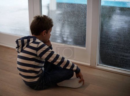 Rainy days with nothing to do...A young boy sitting by the window and looking bored while it rains outside