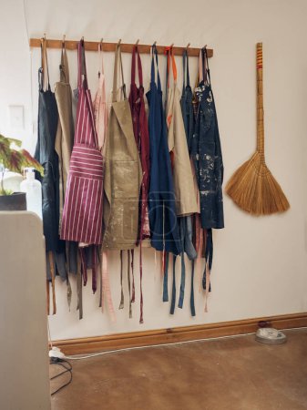 Téléchargez les photos : Clothes rack, apron and pottery with a broom in an empty workshop or studio against still life wall for design. Creative, art and clothing with a group of aprons hanging in a small business startup. - en image libre de droit