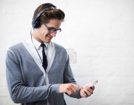 Photo for Downloading more music on his smartphone. A young man smiling at his phone while listening to music on his headphones - Royalty Free Image