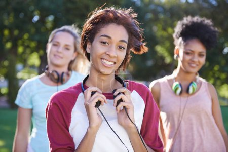 Photo for We love music. Portrait of a smiling young lady standing in a park wearing headphone around her neck with friends standing in the background - Royalty Free Image