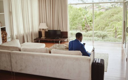 Téléchargez les photos : Phone, travel and hospitality with a business black man sitting on a sofa in the living room of a hotel with his luggage. Back, bag and communication with a male employee typing a text message. - en image libre de droit