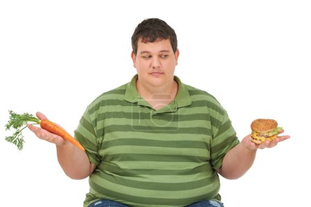 Photo for Tempted to have a taste. An obese young man sitting with a carrot in one hand a burger in the other and peering at the burger with a tempted expression - Royalty Free Image