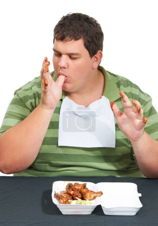 Photo for Fingers full of fat. An obese young man wearing a bib and siting at a table with a container of saucy chicken wings and licking his sauce covered fingers - Royalty Free Image