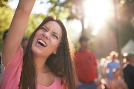 Photo for Feeling the music. A beautiful young woman cheering at a music festival with arm raised in the air and crowd in the background - Royalty Free Image
