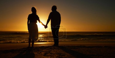 Téléchargez les photos : Silhouette of mature couple holding hands on the beach at sunset. Senior couple bonding on holiday together. Mature couple being affectionate on vacation on the beach. Older couple on seaside vacatio. - en image libre de droit