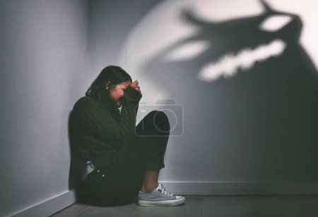 Foto de Fear keeps you from living your best life. a young woman sitting in the corner of a dark room with a scary figure on the wall - Imagen libre de derechos