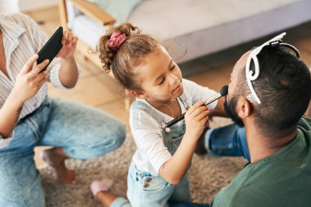 Photo for A little beauty guru in the making. a little girl putting makeup on her father while her mother takes pictures of them at home - Royalty Free Image
