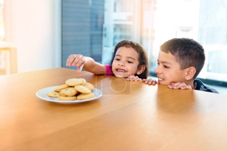 Photo for Almost there, just one more stretch and you have it. two mischievous young children stealing cookies on the kitchen table at home - Royalty Free Image