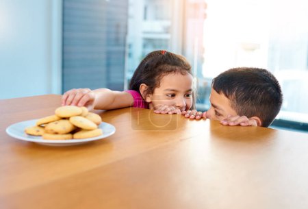 Photo for Dont tell anyone about this okay. two mischievous young children stealing cookies on the kitchen table at home - Royalty Free Image