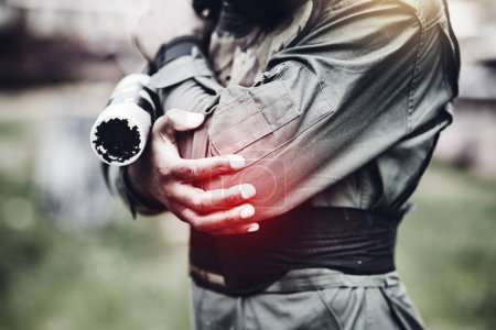 Paintball, pain or man with an elbow injury after playing a shooting game on a fun battlefield on holiday. Red glow, fitness or hands of player in an arm accident in an outdoor military competition.