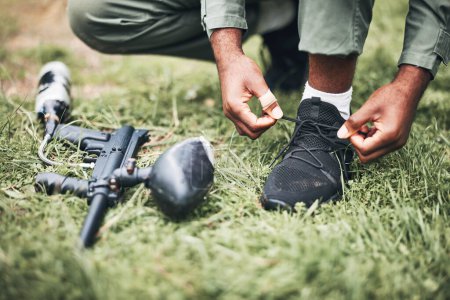 Paintball, gun and shooter or man kneeling ready for competitive match or competition in the forest. Player, athlete and person tying laces with game equipment or weapon on a sports field.