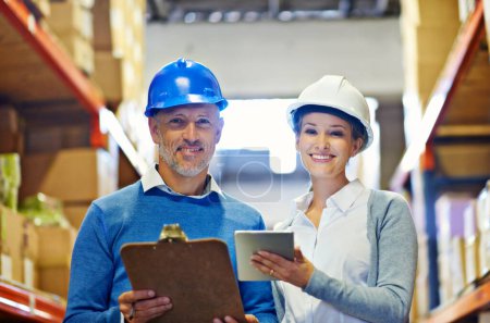Photo for We made a great team. Two people wearing hardhats smiling at the camera in a warehouse - Royalty Free Image
