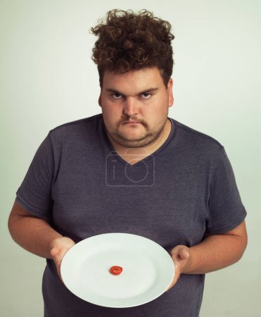 Photo for Wheres the rest of my burger. Portrait of an overweight man holding a plate with a tiny sliver of tomato - Royalty Free Image