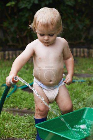 Photo pour Inquisitive Baby. A baby in his nappy holding a hose pipe and pouting water into a gardening container - image libre de droit