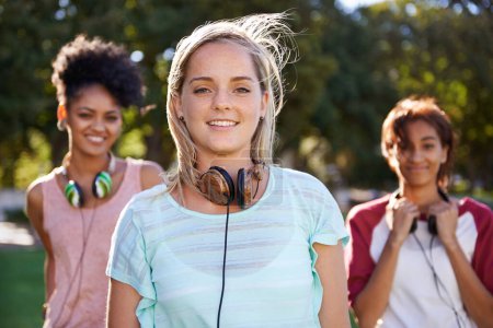Photo for We can listen to the latest tracks where ever we go. Portrait of a smiling young lady standing in a park wearing headphone around her neck with friends standing in the background - Royalty Free Image