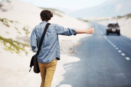 Foto de El viajero aventurero. Vista trasera de un joven de moda haciendo autostop en el lado de la carretera con un coche en la distancia - Imagen libre de derechos