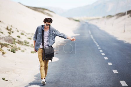 Foto de No todos los que vagan están perdidos. Guapo joven haciendo autostop mientras camina por el lado de la carretera - Imagen libre de derechos