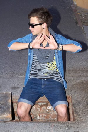 Foto de Going down. A stylishly-dressed young man partially sitting in a hole in the pavement and making a hand gesture - Imagen libre de derechos