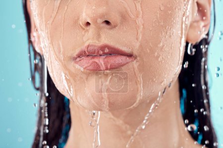 Foto de Water splash, skincare and lips of woman in shower in studio isolated on a blue background. Beauty, face and young female model washing, cleaning or bathing for hygiene, wellness and healthy skin - Imagen libre de derechos