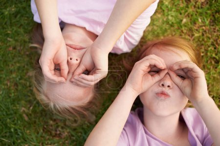 Téléchargez les photos : On peut te voir. Deux petites filles allongées sur le dos faisant des cercles autour de leurs yeux avec leurs mains - en image libre de droit