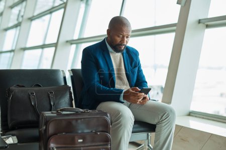 Photo for Black man, phone and luggage at airport for business travel, trip or communication waiting for flight. African American male traveler chatting or checking plain times, schedule or delay on smartphone. - Royalty Free Image