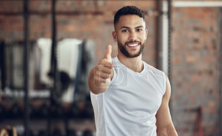 Téléchargez les photos : Portrait of young athlete giving thumbs up. Young fit man doing his best to endorse exercise in the gym. Young athlete making a gesture in the gym. Muscular young man making a hand gesture in the gym. - en image libre de droit