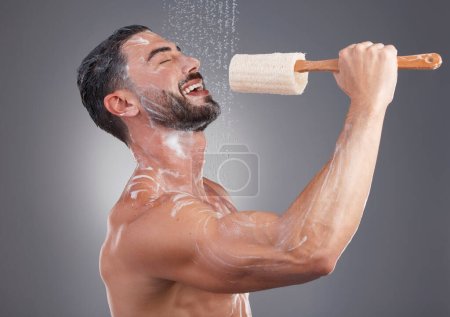 Téléchargez les photos : Shower, cleaning and man singing with brush in studio for skincare, washing body and wellness on gray background. Beauty, bathroom and happy male do karaoke with loofah sponge, soap and water splash. - en image libre de droit