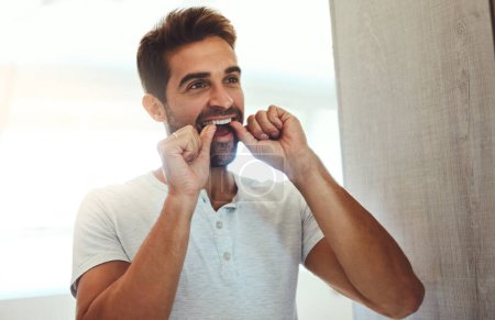 Téléchargez les photos : Getting in to those hard to reach places. a handsome young man flossing his teeth while looking at his reflection in the mirror at home - en image libre de droit
