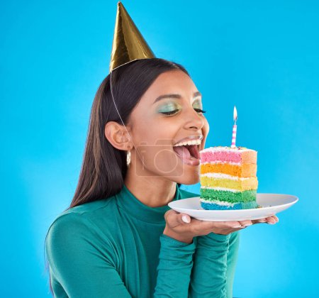 Celebrate, birthday and woman in studio with a cake, party hat and candle for a celebration. Happy, smile and Indian female model ready to eat sweet rainbow dessert while isolated by blue background