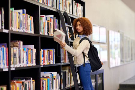 Téléchargez les photos : Elle a trouvé ce qu'elle cherchait. une jeune femme étudiant à partir d'un livre tout en se tenant près d'une bibliothèque - en image libre de droit