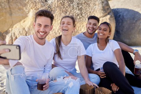 Ein paar Freunde, Selfie und Drinks bei einem Strandpicknick mit einem Lächeln für ein Social-Media-Update während des Urlaubs im Sommer. Männer und Frauen mit Handy bei doppeltem Date an Felsen für Spaß und Freiheit im Freien.