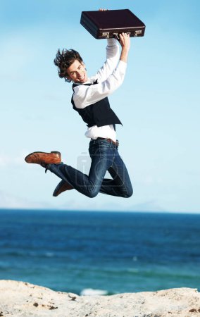 Foto de La libertad de ser autónomo. Feliz joven empresario saltando sosteniendo su maletín en el aire con el océano en el fondo - Imagen libre de derechos
