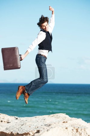 Foto de Es genial estar fuera de la oficina. Joven sosteniendo un maletín y saltando en el aire con el océano en el fondo - Imagen libre de derechos