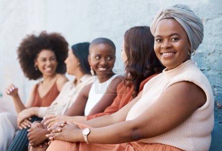 Foto de Joven mujer de negocios afroamericana sentada en una fila con sus compañeros de trabajo contra una pared fuera de la ciudad. Negro hembra sonriendo mientras se sienta al aire libre con sus colegas. - Imagen libre de derechos