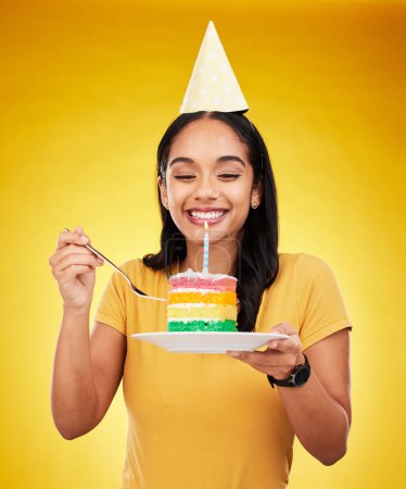 Woman is eating cake, birthday celebration and happy in portrait, rainbow dessert and candle on yellow background. Celebrate, festive and young female, excited for sweet treat and party hat in studio.