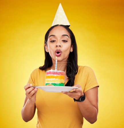 Woman, birthday cake and celebration, blow out candle with rainbow dessert isolated on yellow background. Celebrate, festive and young female, making a wish with sweet treat and party hat in studio.