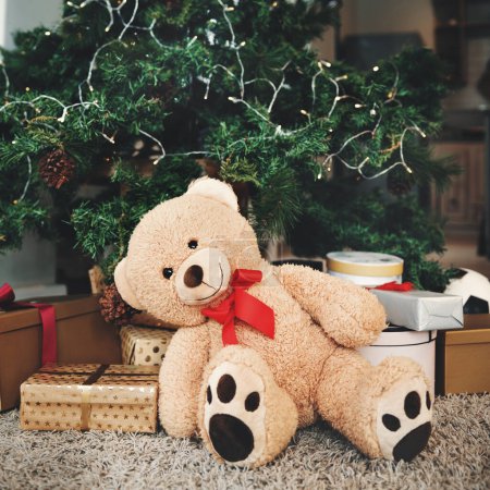 Téléchargez les photos : Noël, cadeau et fête avec un ours en peluche près d'un arbre, prêt pour la célébration pendant la période des fêtes. Décembre, événement et un animal en peluche assis dans le salon d'un appartement en cadeau. - en image libre de droit