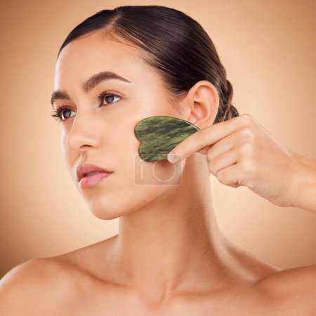 Beauty, gua sha and face of woman for skincare, facial treatment and wellness with spa stone. Salon, dermatology and girl on brown background with cosmetics, face massage and luxury tools in studio.